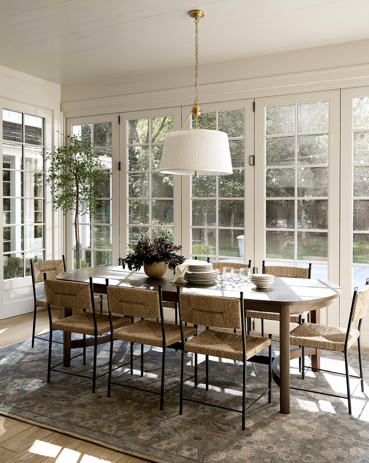 A bright dining room showcases the Gardiner Dining Table with eight woven chairs on a patterned rug, illuminated by large windows. A potted plant and chandelier complete the space, by McGee & Co.