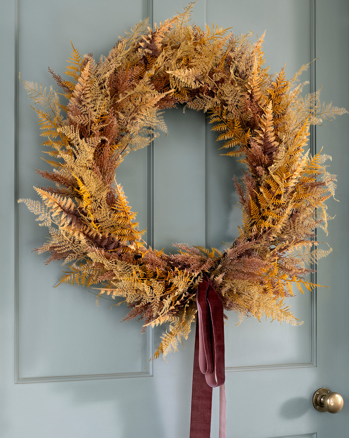 The 30 Faux Golden Fern Wreath features dried golden-brown ferns and a velvet burgundy ribbon, hanging on a light blue door with a brass round doorknob—an elegant fall wreath that adds timeless entryway charm, by McGee & Co.