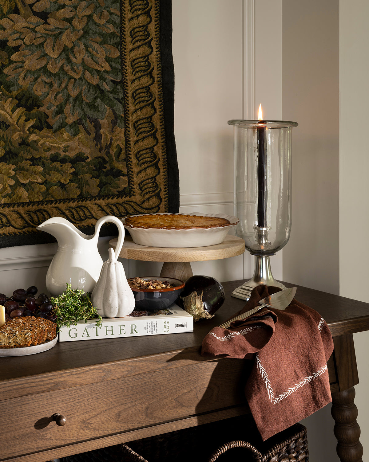 A wooden table displays a pie, a candle in a glass holder, a white pitcher, a ceramic pear, kitchen accessories including the Broadleaf Tea Towel, plus grapes and nuts in a bowl, eggplant, brown napkin, and the book Gather beneath a tapestry by McGee & Co.