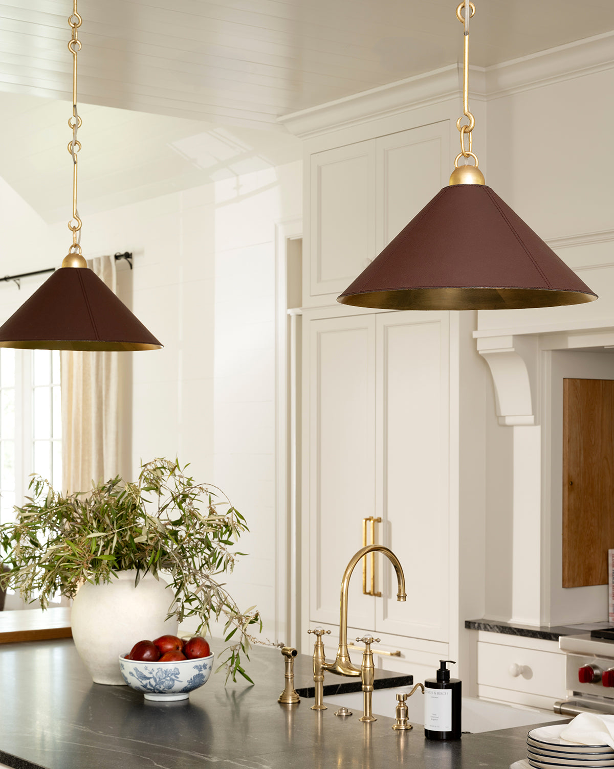 A bright kitchen featuring brass fixtures, a black countertop, elegant lighting from two Scarlett Pendants in brown and gold, a white vase with greenery, apples in a bowl, white cabinetry, and a large window by McGee & Co.