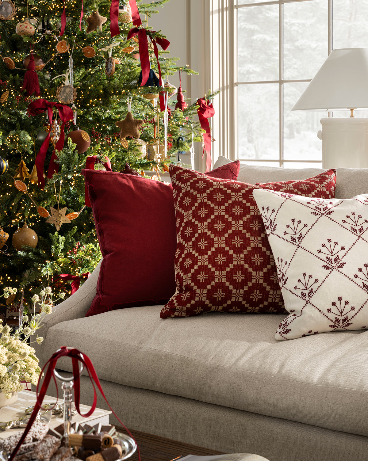 A beige sofa with McCallister Pillow Covers and red-and-white patterned pillows sits by a festive Christmas tree. Sunlight fills the room, highlighting holiday decor and treats arranged on the table in front, by McGee & Co.