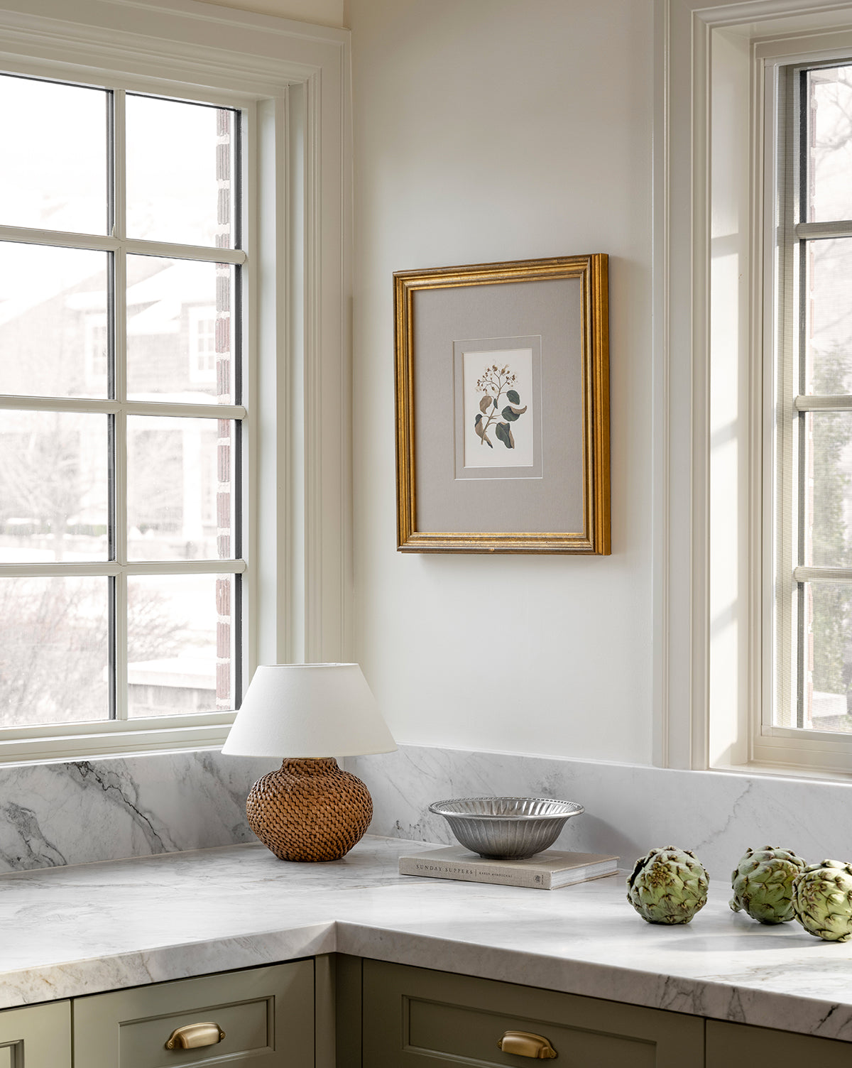 A bright kitchen corner features large windows, a marble countertop, green cabinets, a wicker lamp with a white shade, a silver bowl on books, two artichokes, and the Blossoming Branch vintage wall art in an antique gold frame, by McGee & Co.