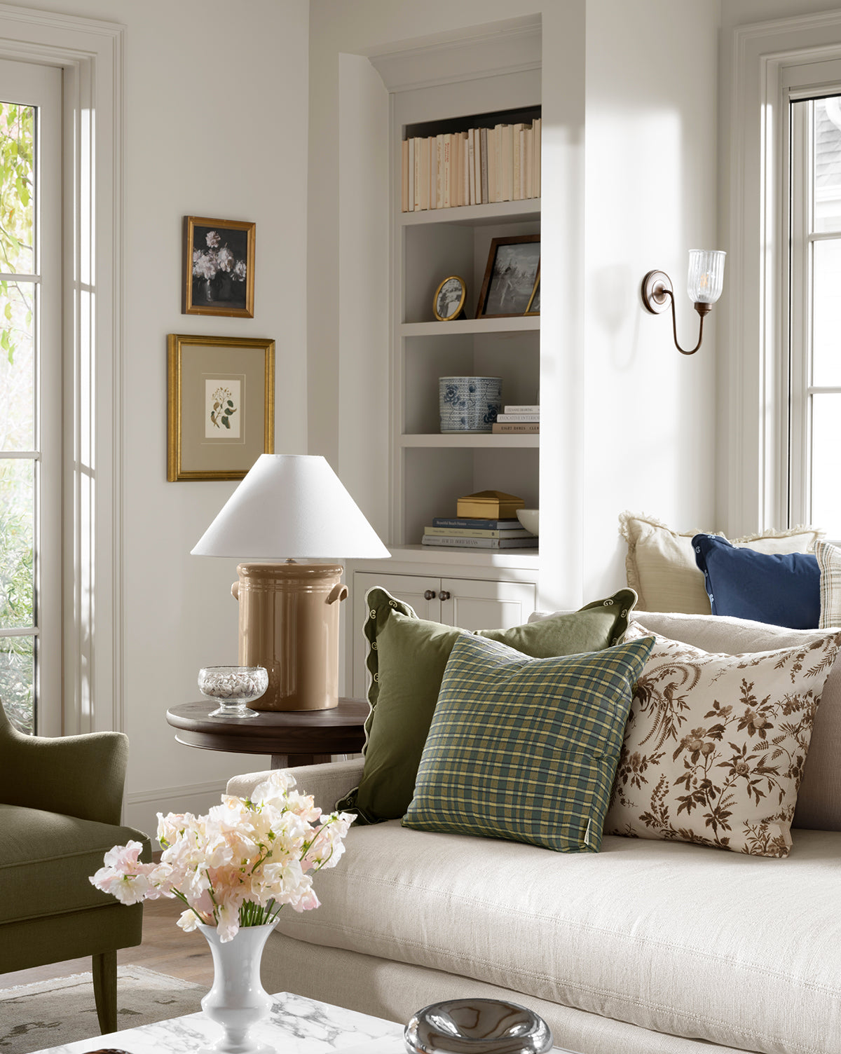 A bright living room features a beige sofa, timeless accent pillows including the Selwyn Plaid Pillow Cover, a side table with a lamp, a marble coffee table topped with pink flowers, built-in shelves of books and decor, and sunlight from large windows, by McGee & Co.
